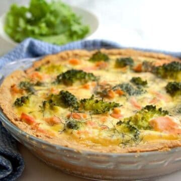 A freshly baked salmon and broccoli quiche in a glass pie dish, served on a white table with a lettuce salad in the background.