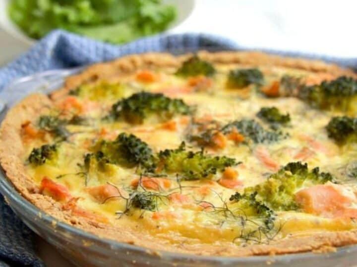 A freshly baked salmon and broccoli quiche in a glass pie dish, served on a white table with a lettuce salad in the background.