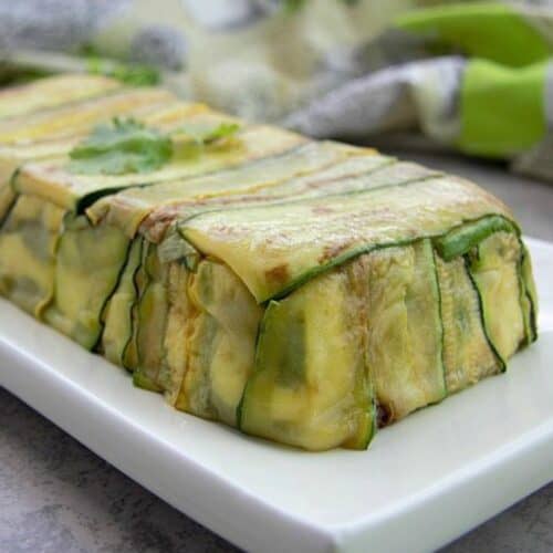 A zucchini terrine is presented on a white plate with a sprig of cilantro on top.