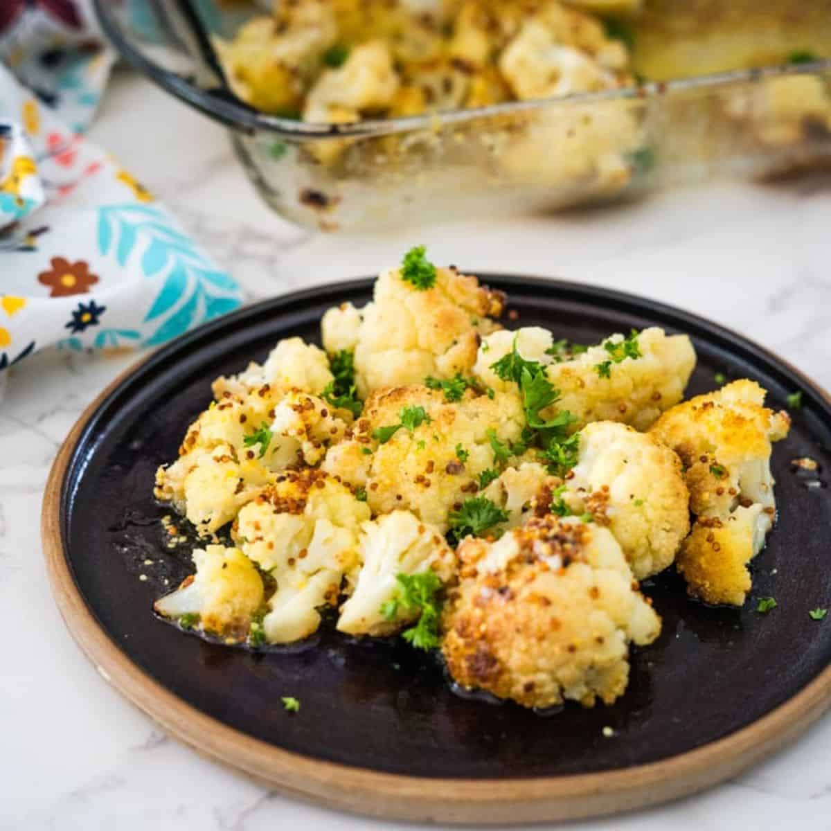 Keto roasted cauliflower on a black plate, garnished with parsley, with a glass baking dish in the background.