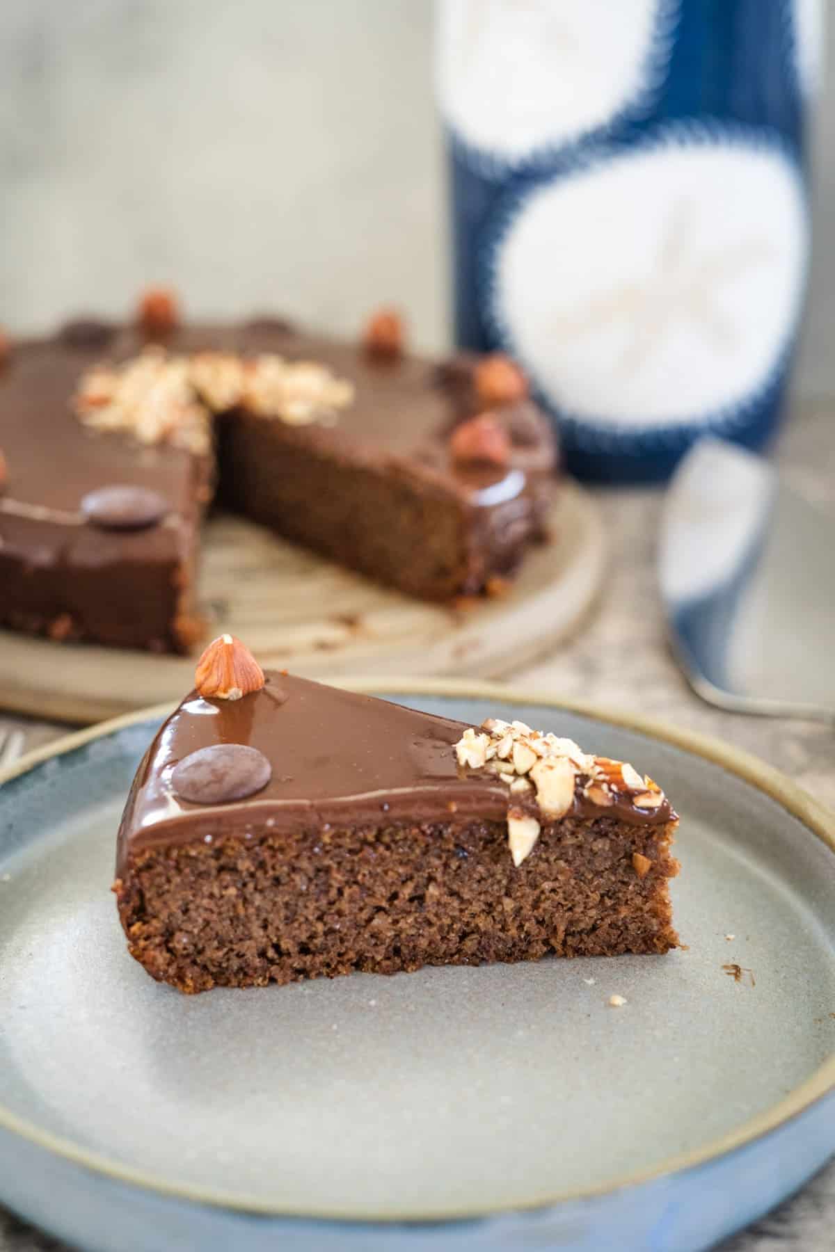 A slice of decadent chocolate hazelnut cake topped with a whole hazelnut and chopped nuts rests on a plate; the whole cake with additional slices is tempting in the background.