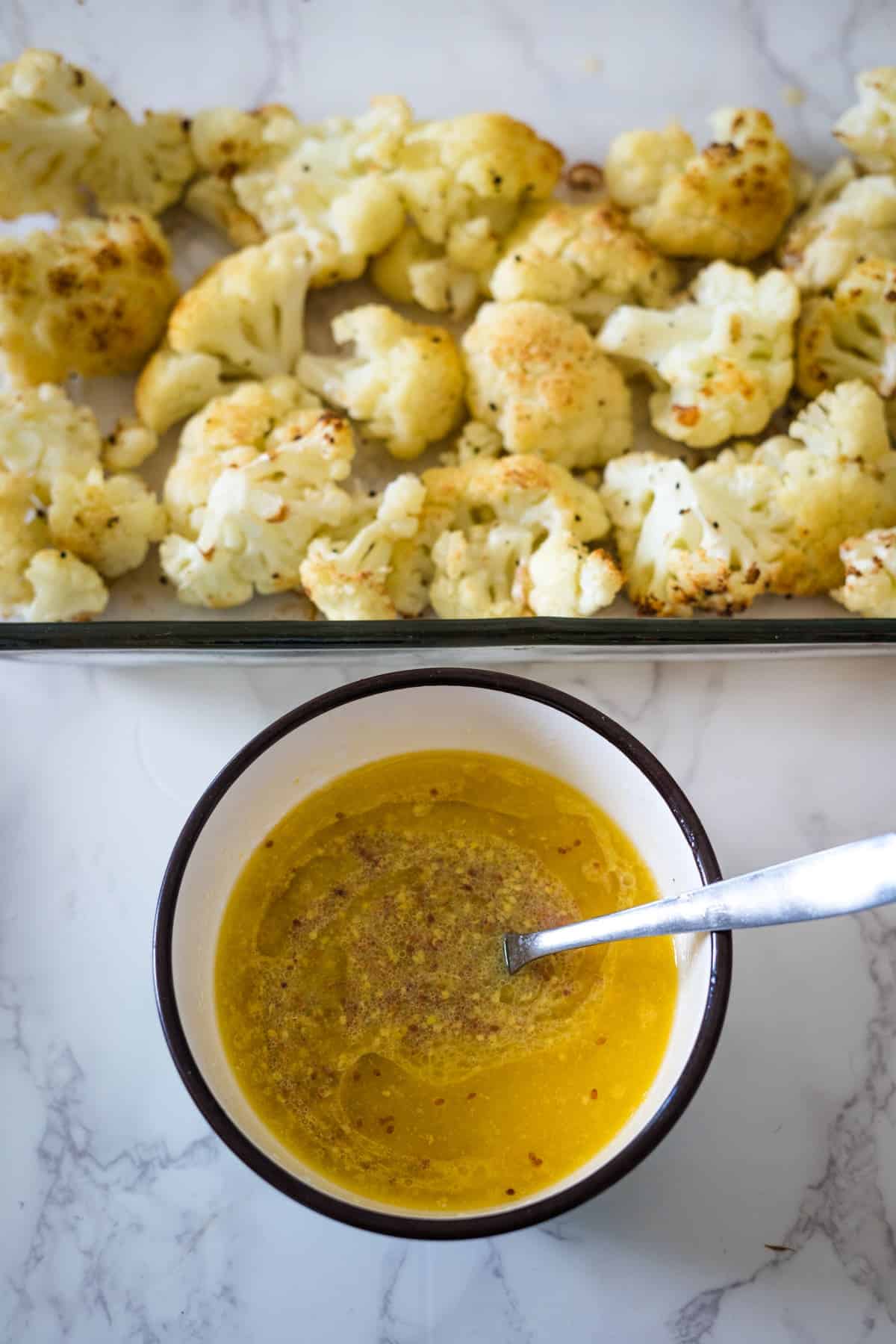 A bowl of yellow sauce with a spoon, next to a tray of keto roasted cauliflower on a marble countertop.