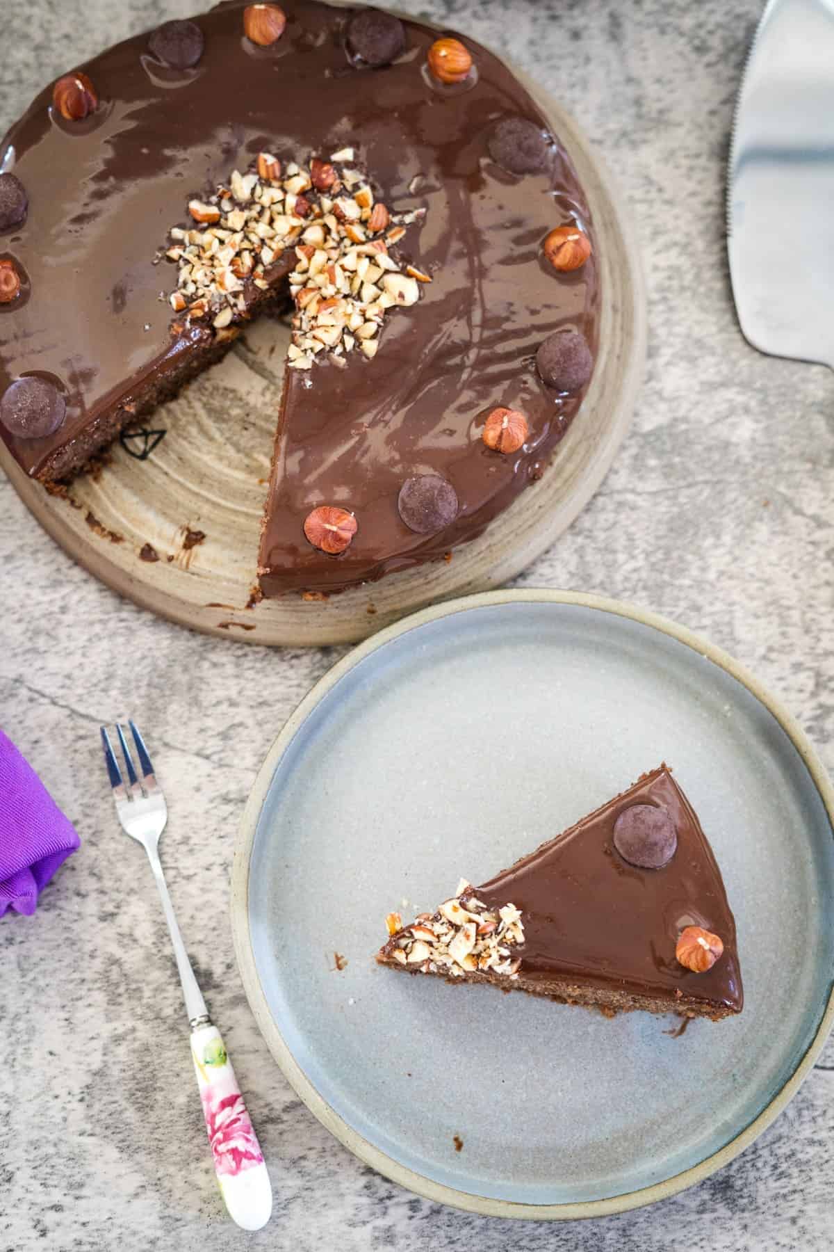 A slice of chocolate hazelnut torte topped with chopped nuts and chocolate drops sits on a separate plate, alongside a fork with a floral handle on a grey surface.