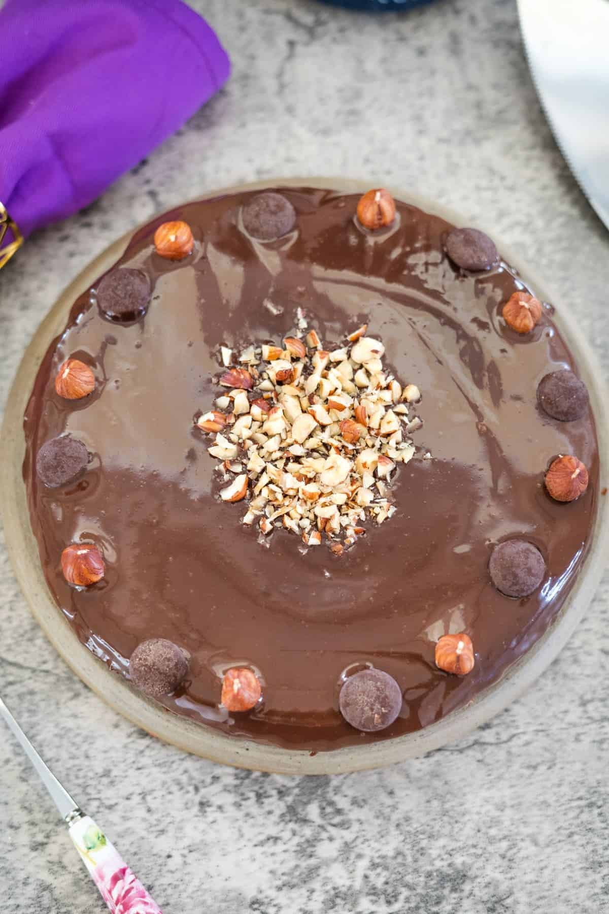 A round chocolate hazelnut cake with a glossy surface, topped with chopped nuts in the center, chocolate buttons, and whole hazelnuts around the edge.