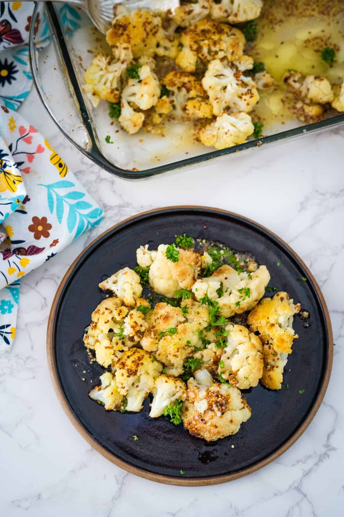 Roasted keto cauliflower on a black plate, garnished with herbs. A baking dish with more cauliflower sits in the background.