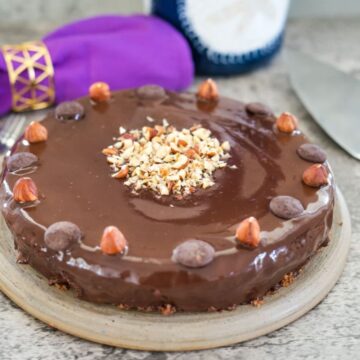 Round chocolate hazelnut cake garnished with whole and chopped hazelnuts on a plate, with a purple napkin and a fork in the background.