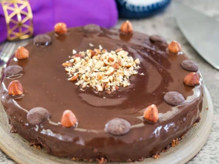 Round chocolate hazelnut cake garnished with whole and chopped hazelnuts on a plate, with a purple napkin and a fork in the background.