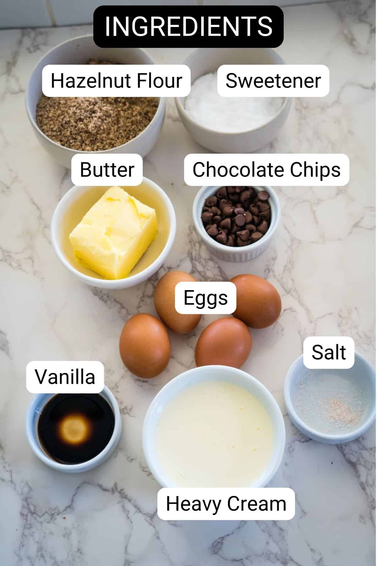 Top view of nine baking ingredients in bowls on a marble surface labeled: hazelnut flour, sweetener, butter, chocolate chips, eggs, salt, vanilla, heavy cream. Text above reads "INGREDIENTS." Perfect for a chocolate hazelnut treat!