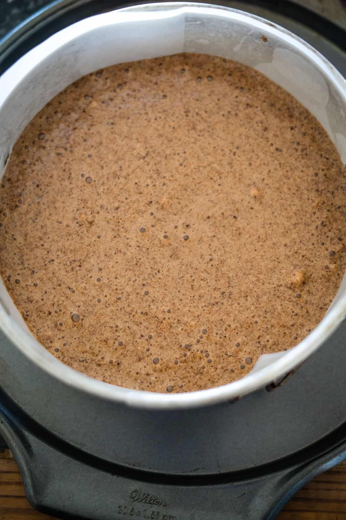 A round cake pan filled with uncooked batter, lined with parchment paper, sits on a black countertop, ready to become a delicious chocolate hazelnut delight.