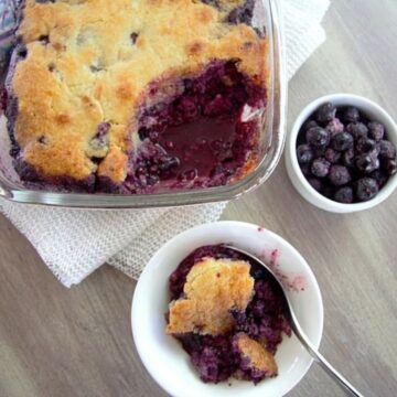 A glass baking dish and a bowl containing freshly baked keto berry cobbler with a spoon, served on a wooden table, with a small bowl of whole blueberries beside it.