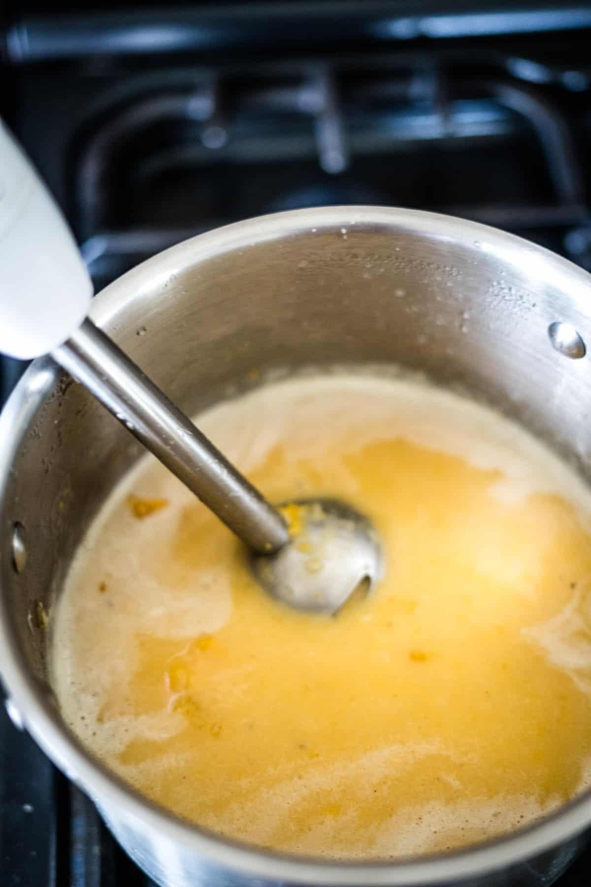 A hand blender is immersed in a pot of pumpkin ginger soup on a stovetop.