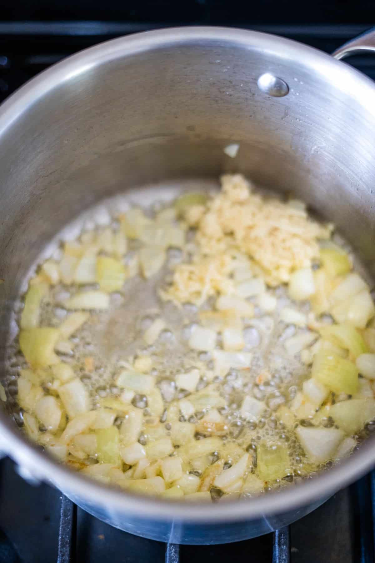 A saucepan on a stove contains chopped onions sautéing in oil, with minced garlic and a hint of ginger added to enhance the flavor.