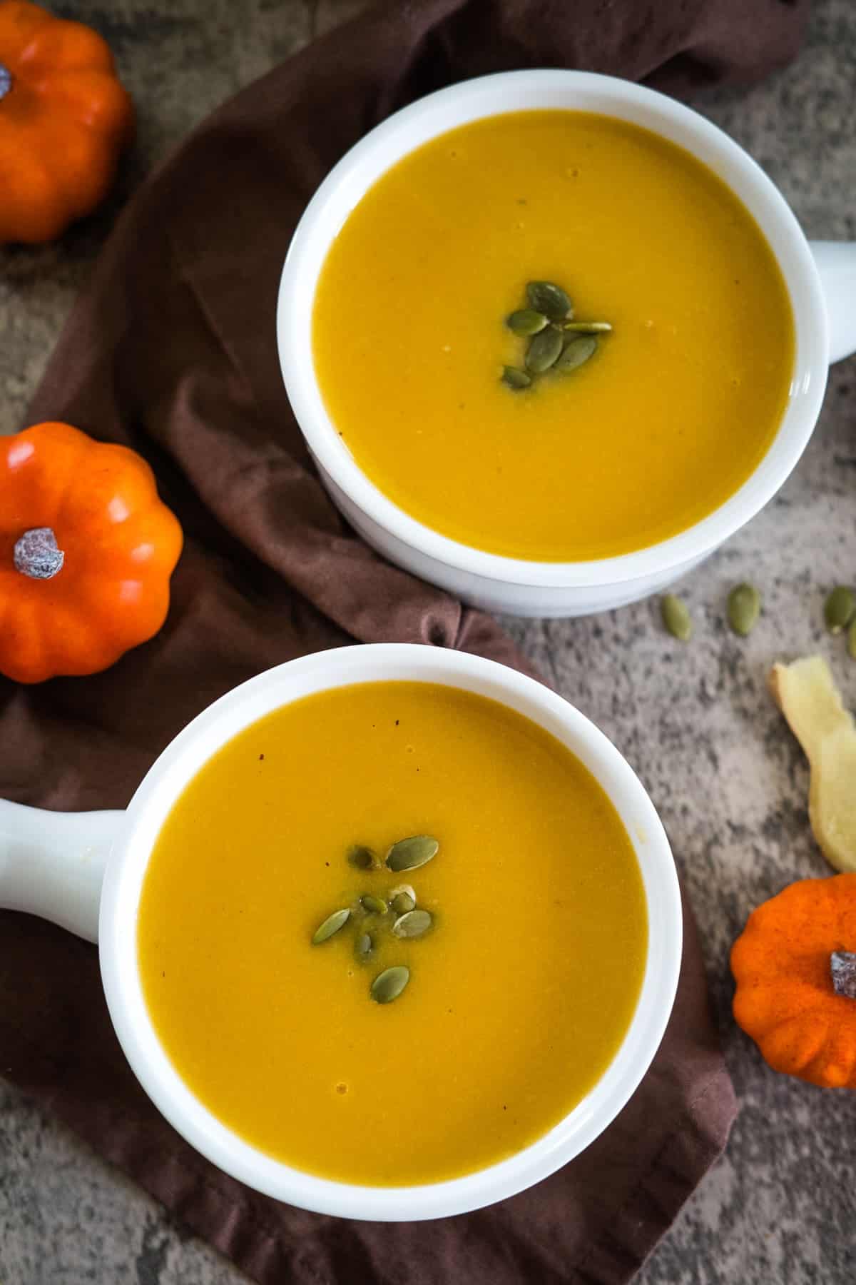 Two white bowls of pumpkin ginger soup garnished with seeds are placed on a dark surface with small orange pumpkins and a brown napkin.