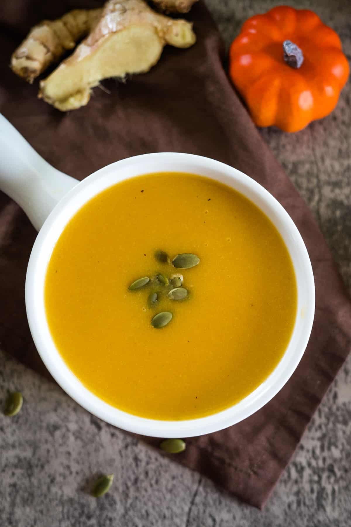 A bowl of pumpkin ginger soup topped with pumpkin seeds sits on a brown cloth. Ginger and a small pumpkin are in the background.