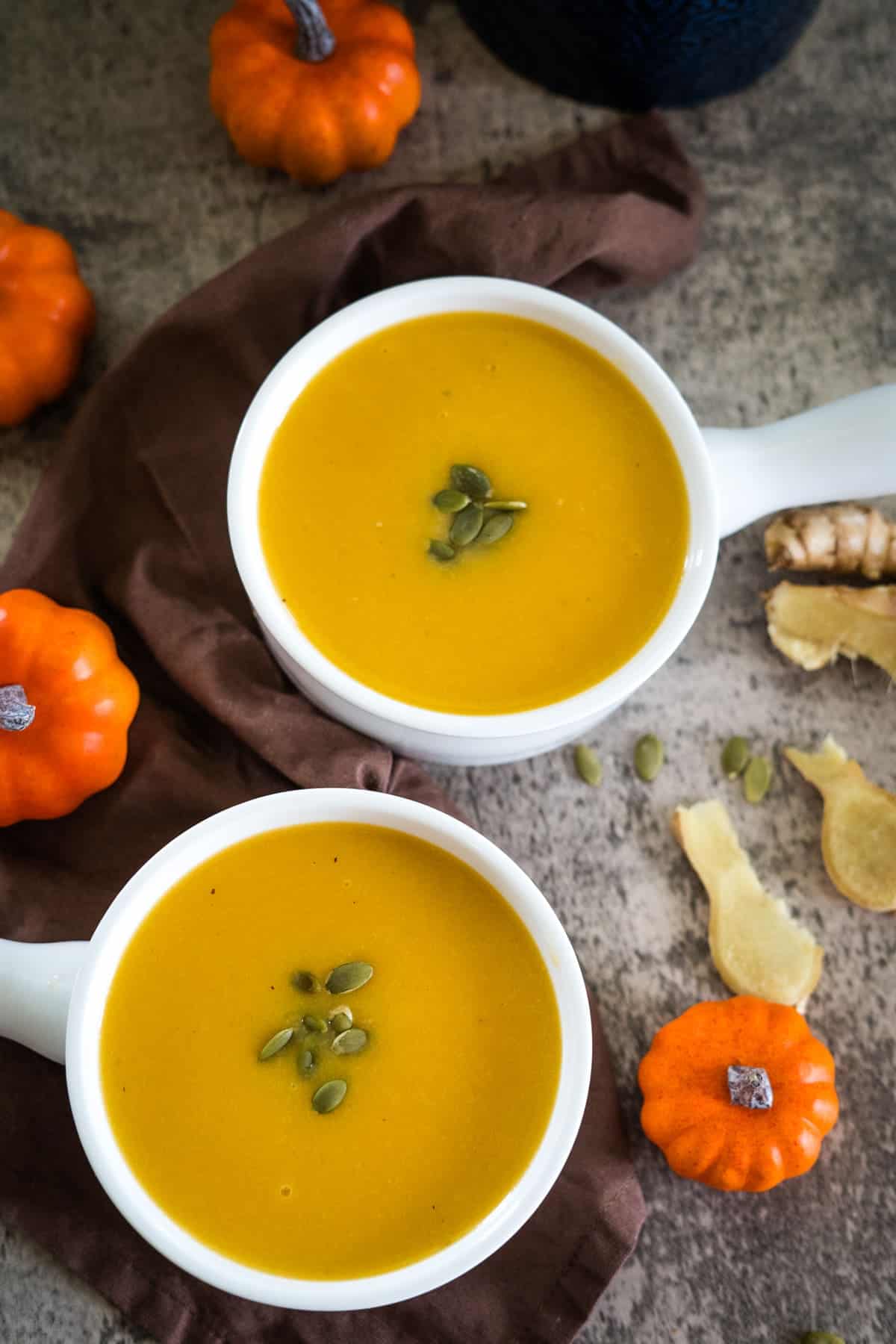 Two white bowls of pumpkin ginger soup garnished with pumpkin seeds, surrounded by small pumpkins, ginger slices, and a brown cloth.