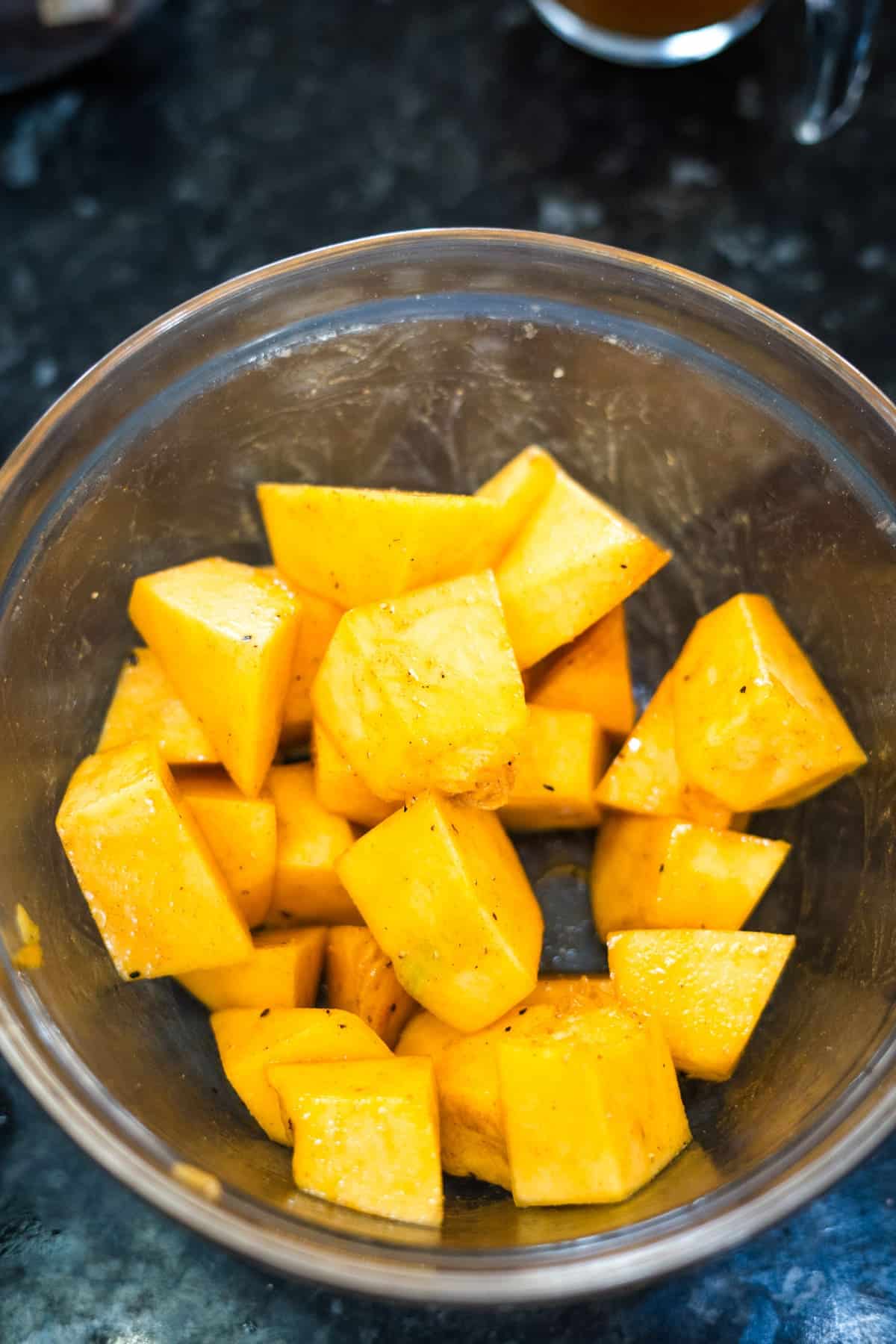 A glass bowl filled with large, evenly cut chunks of raw yellow squash and a hint of pumpkin ginger on a dark countertop.