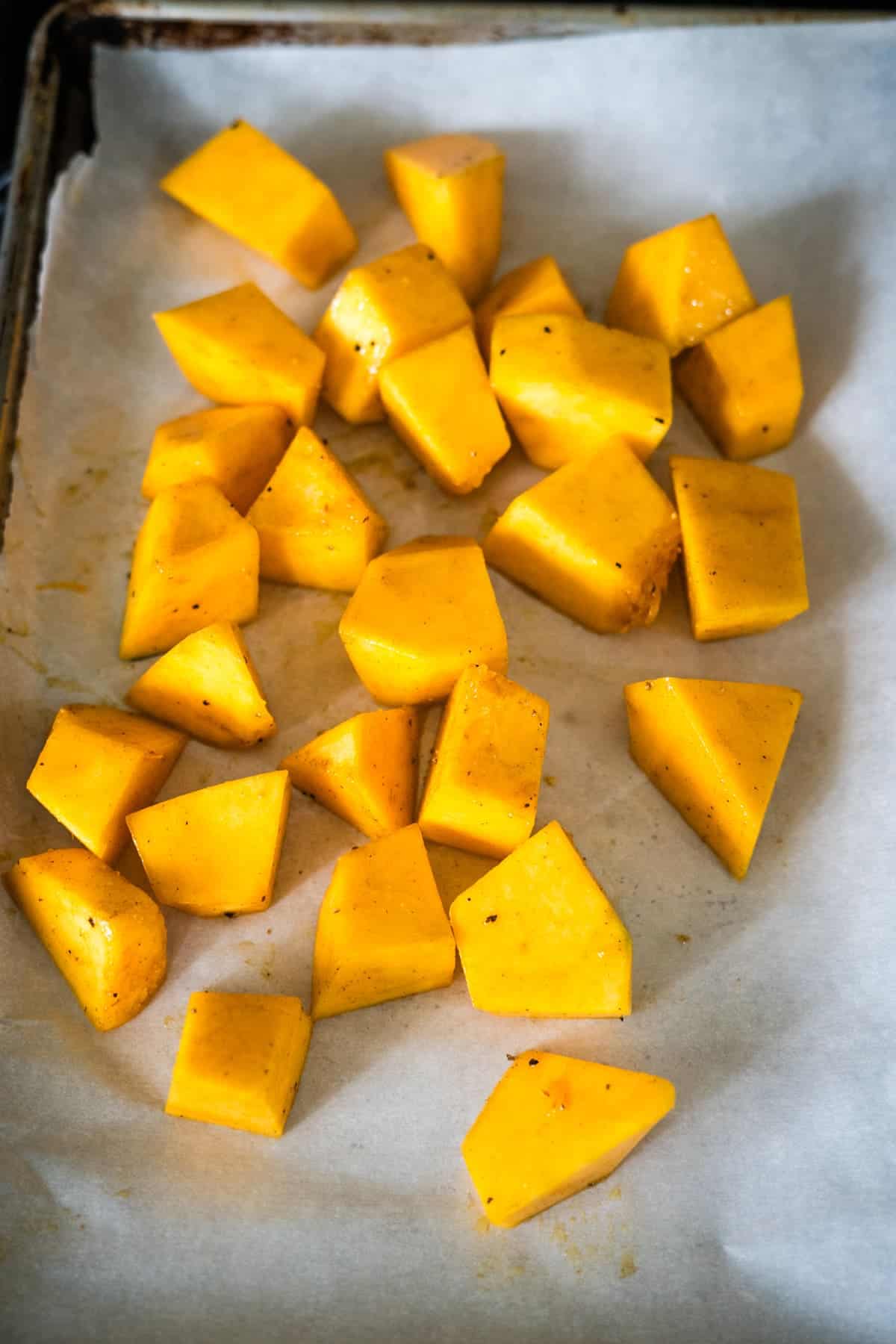 Cubed pieces of butternut squash, with a hint of pumpkin spice and ginger, are spread out on a parchment-lined baking sheet, ready for roasting.