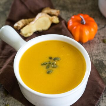 A bowl of pumpkin ginger soup garnished with pumpkin seeds, placed on a brown cloth beside ginger slices and a small decorative pumpkin.