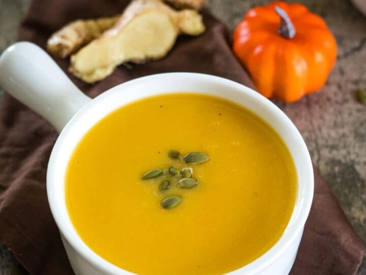 A bowl of pumpkin ginger soup garnished with pumpkin seeds, placed on a brown cloth beside ginger slices and a small decorative pumpkin.