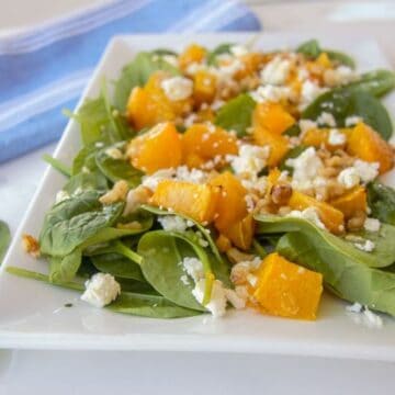 A plate of spinach salad topped with roasted pumpkin, walnuts, and crumbled feta cheese, served on a white square plate with a blue-striped napkin in the background.