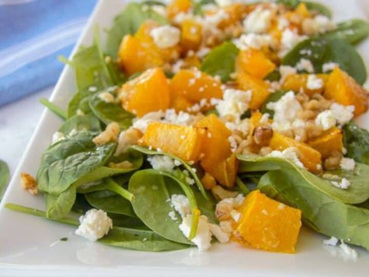 A plate of spinach salad topped with roasted pumpkin, walnuts, and crumbled feta cheese, served on a white square plate with a blue-striped napkin in the background.