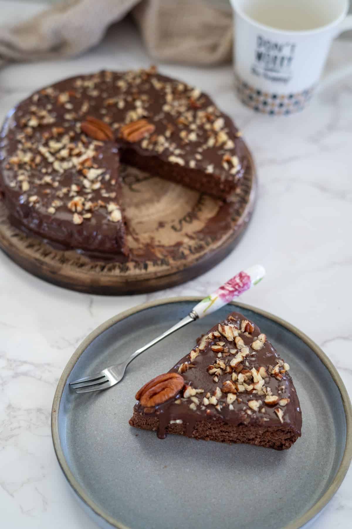 A slice of chocolate pecan torte topped with nuts and a pecan sits on a gray plate next to a fork. The rest of the cake, partially sliced, rests enticingly on a wooden board in the background.