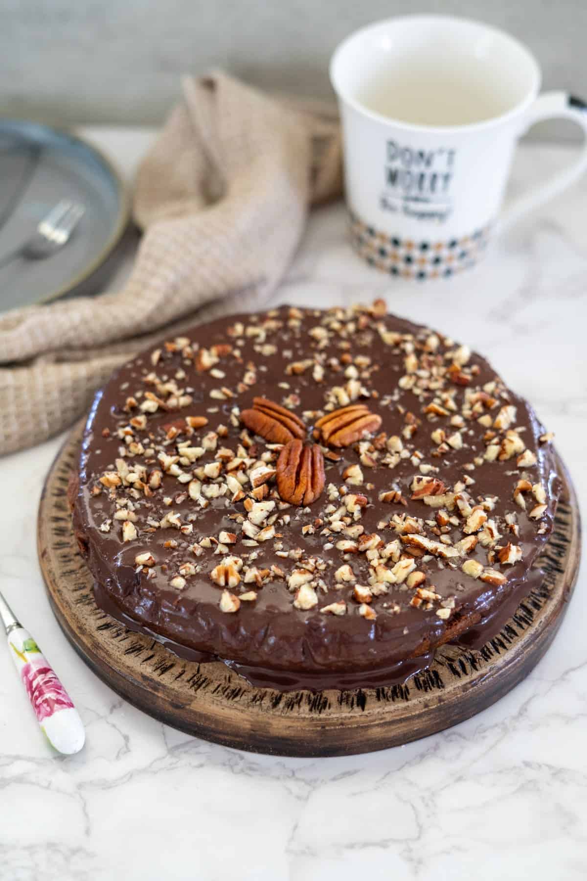 A decadent pecan torte, topped with chopped nuts and rich chocolate, rests on a wooden plate. It's accompanied by a mug and a knife elegantly placed on the cool marble surface.