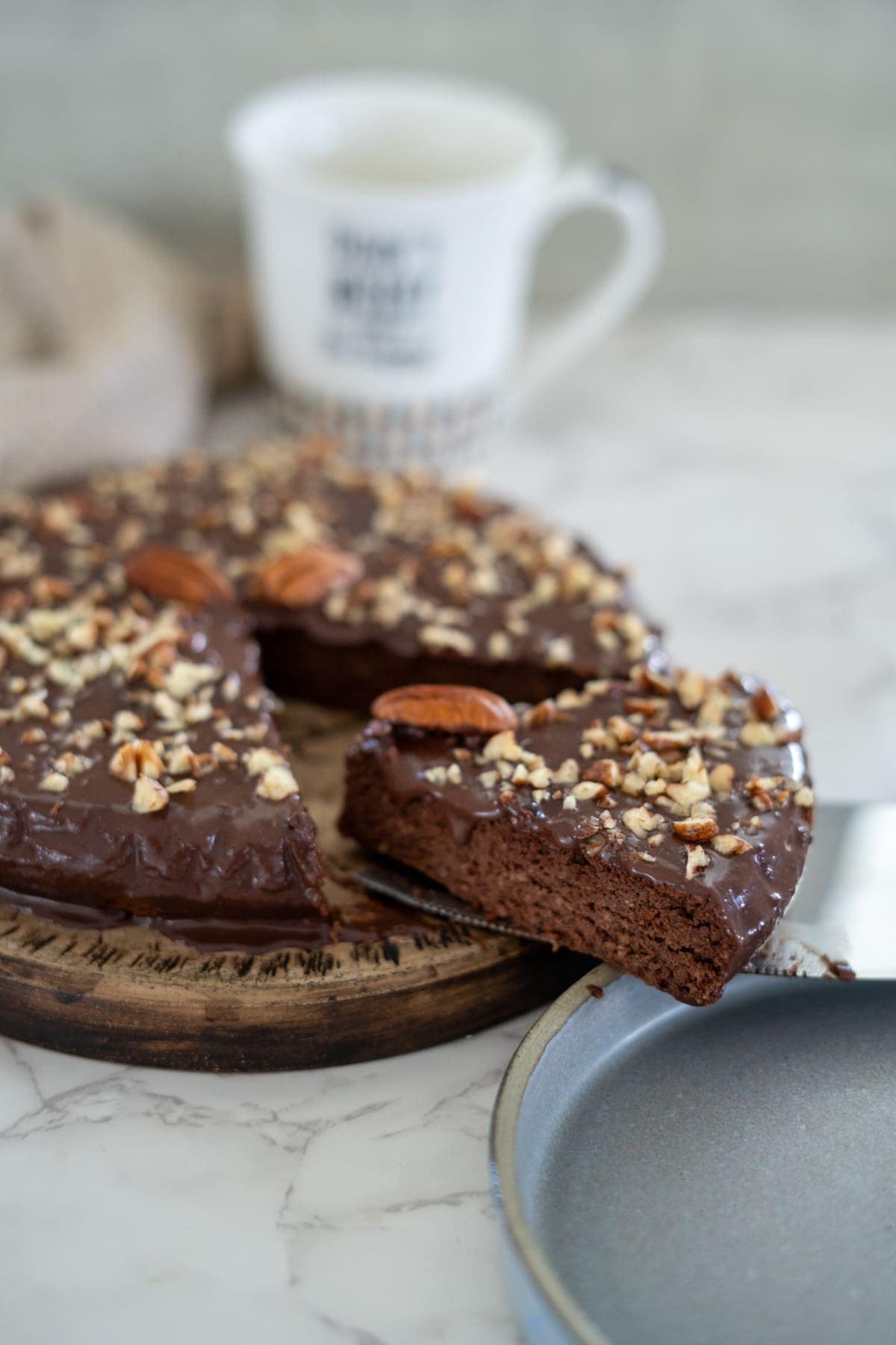 A pecan torte topped with chopped nuts and one slice resting on a metal spatula. A mug is blurred in the background.