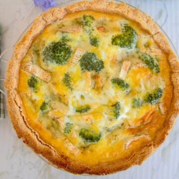 A baked broccoli quiche with a golden-brown crust, filled with melted cheese and eggs, displayed on a white marble surface.