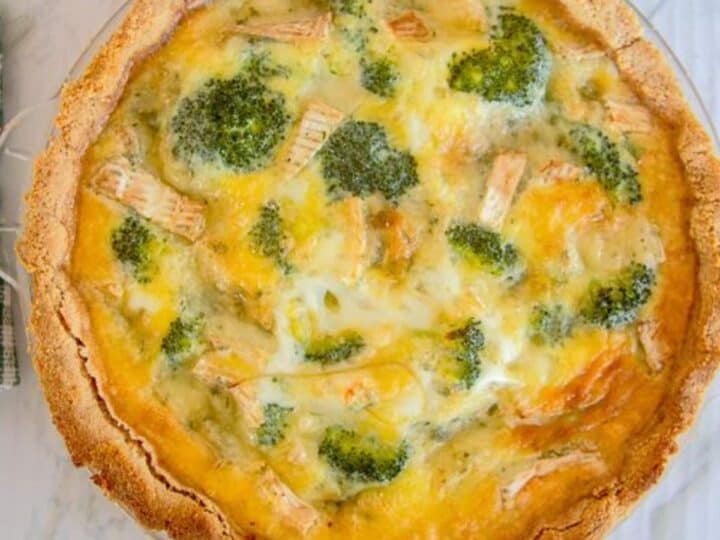 A baked broccoli quiche with a golden-brown crust, filled with melted cheese and eggs, displayed on a white marble surface.