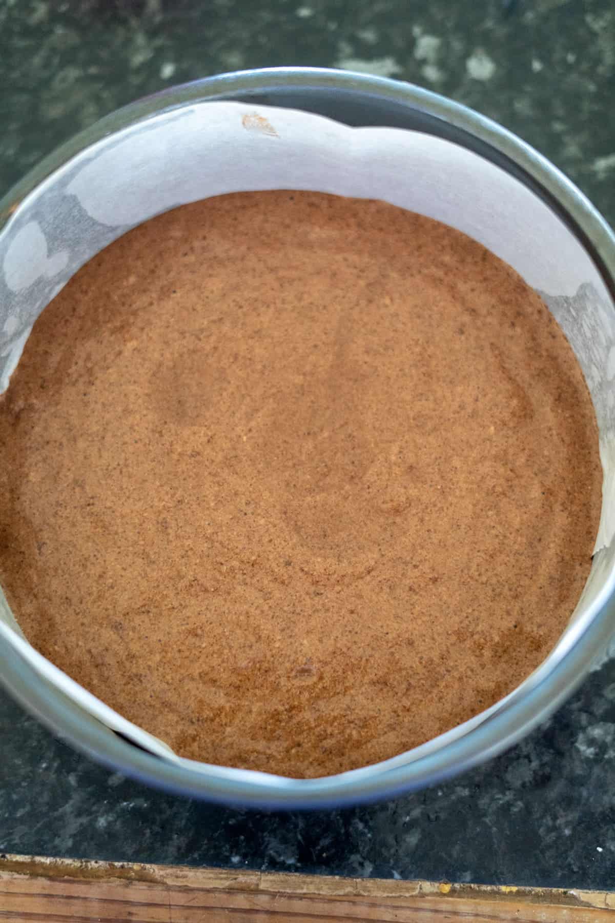 Brown batter for a pecan torte rests in a round baking tin lined with parchment paper on a dark countertop.