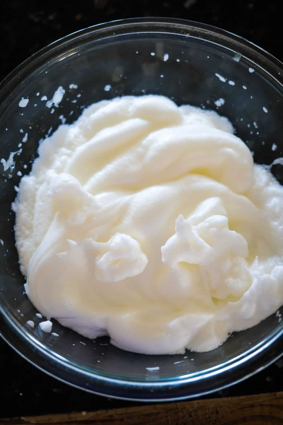 A glass bowl containing whipped egg whites, ready to craft the perfect pecan torte, sits on a dark countertop.