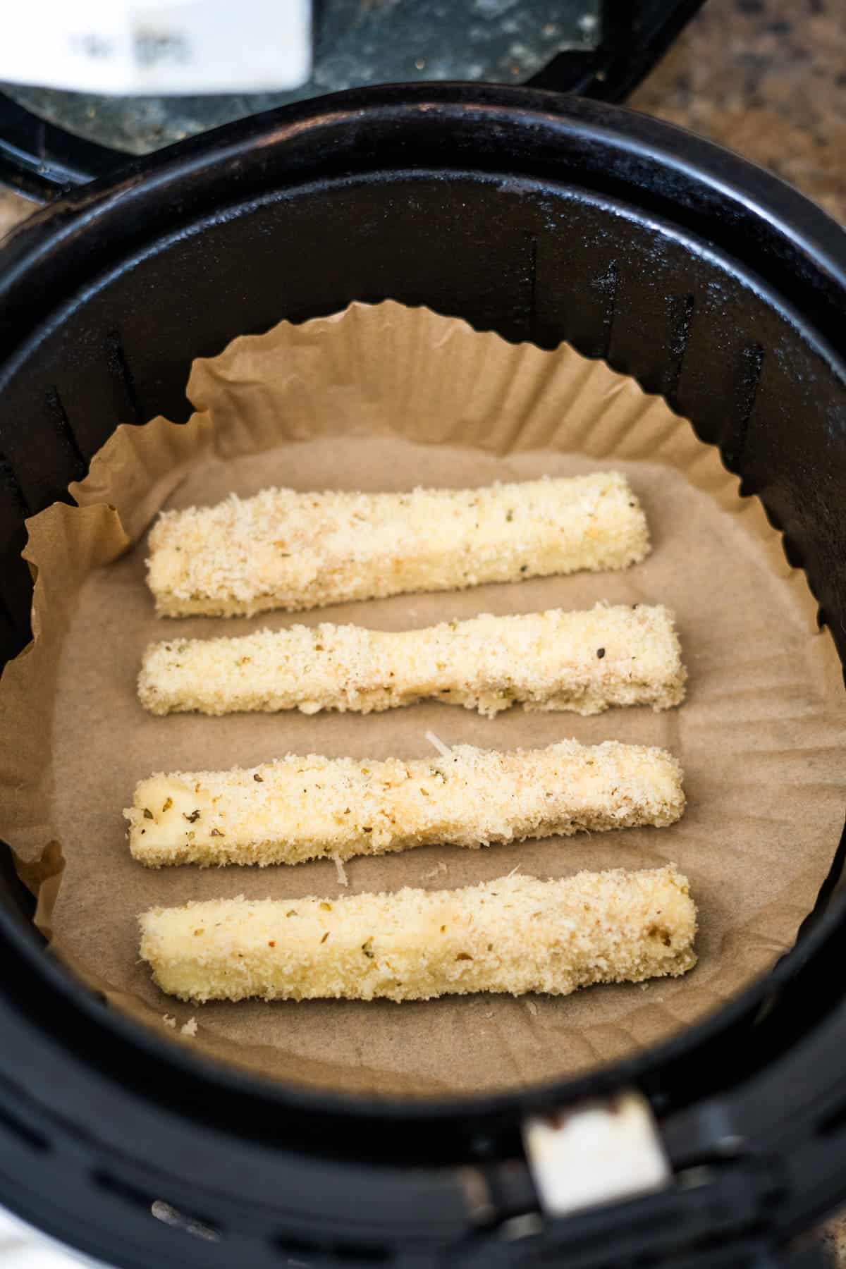 Four breaded mozzarella sticks are placed on parchment paper inside an air fryer basket, alongside a handful of eggplant fries, ready to be cooked.