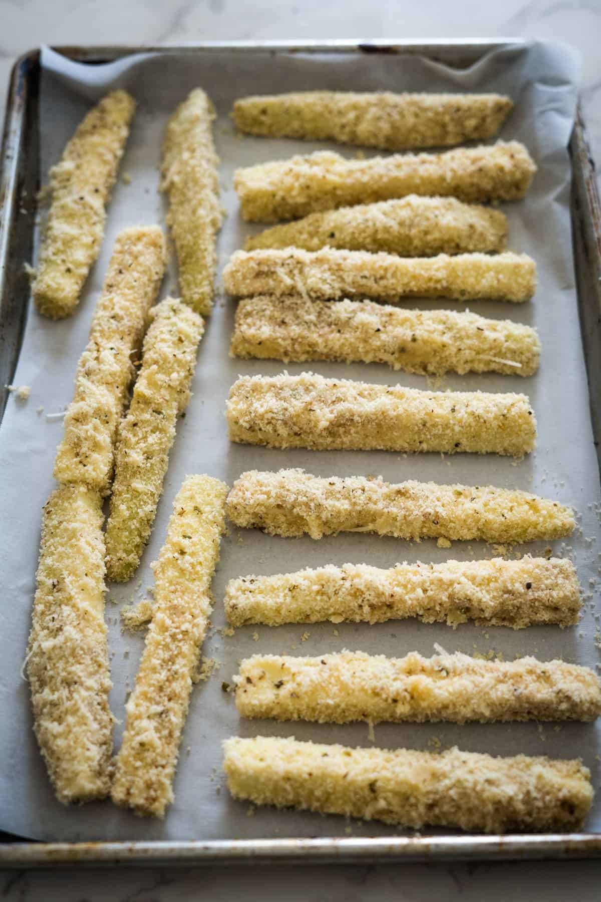 Breaded zucchini sticks and eggplant fries are arranged in rows on a parchment-lined baking sheet.