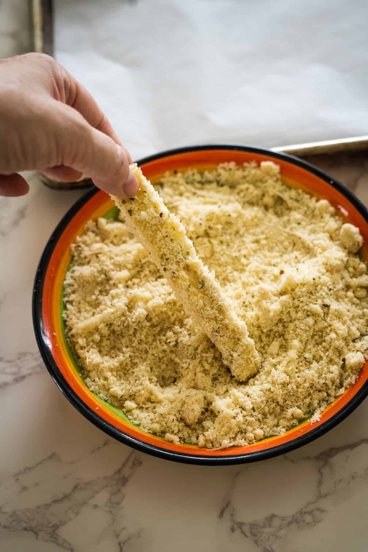 A hand is dipping eggplant fries into a bowl of bread crumbs on a marble countertop.