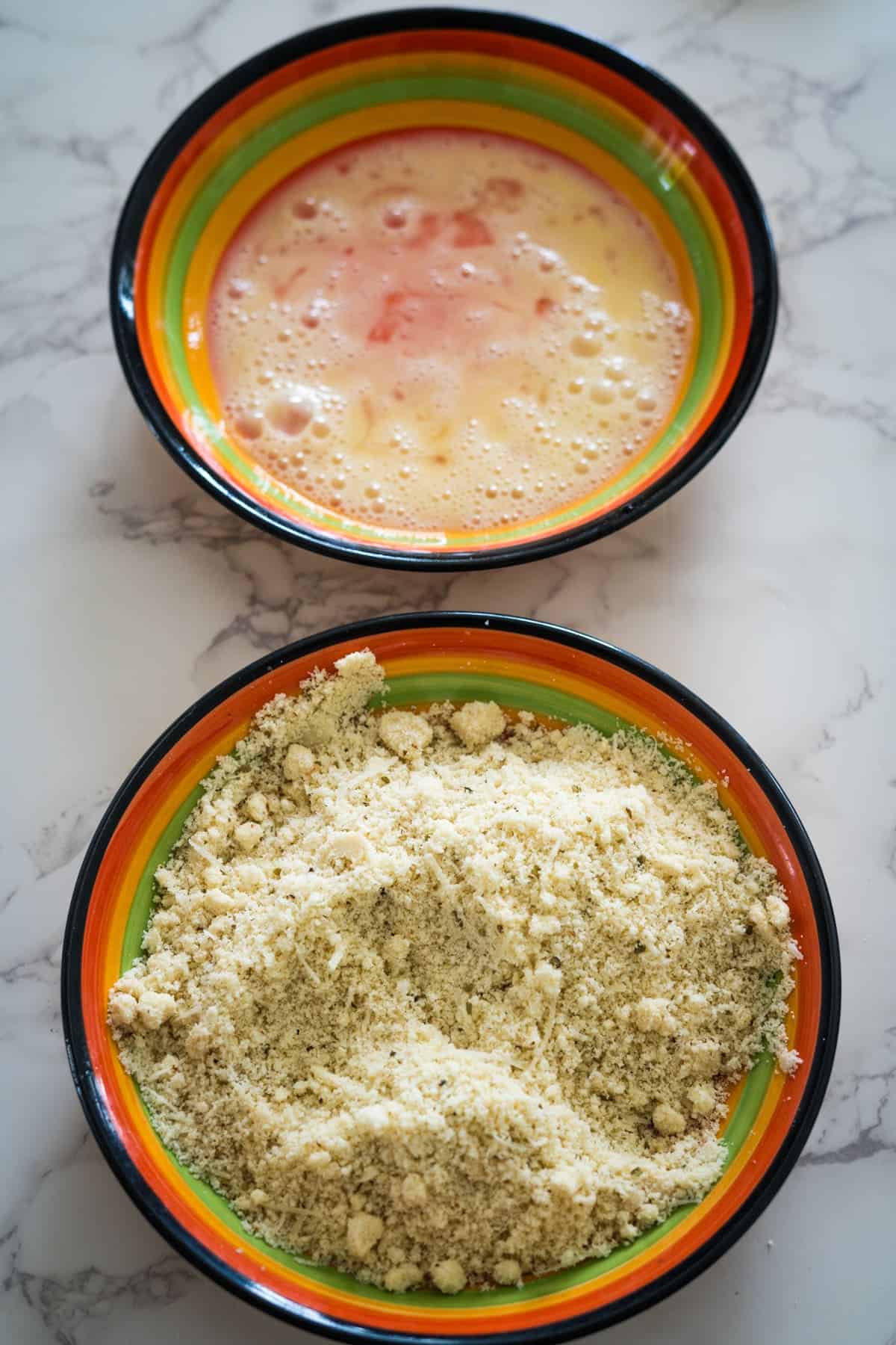 Two bowls on a marble surface; the top bowl contains a mixed egg wash, and the bottom bowl holds a breadcrumb mixture with seasonings, ready for making crispy eggplant fries.