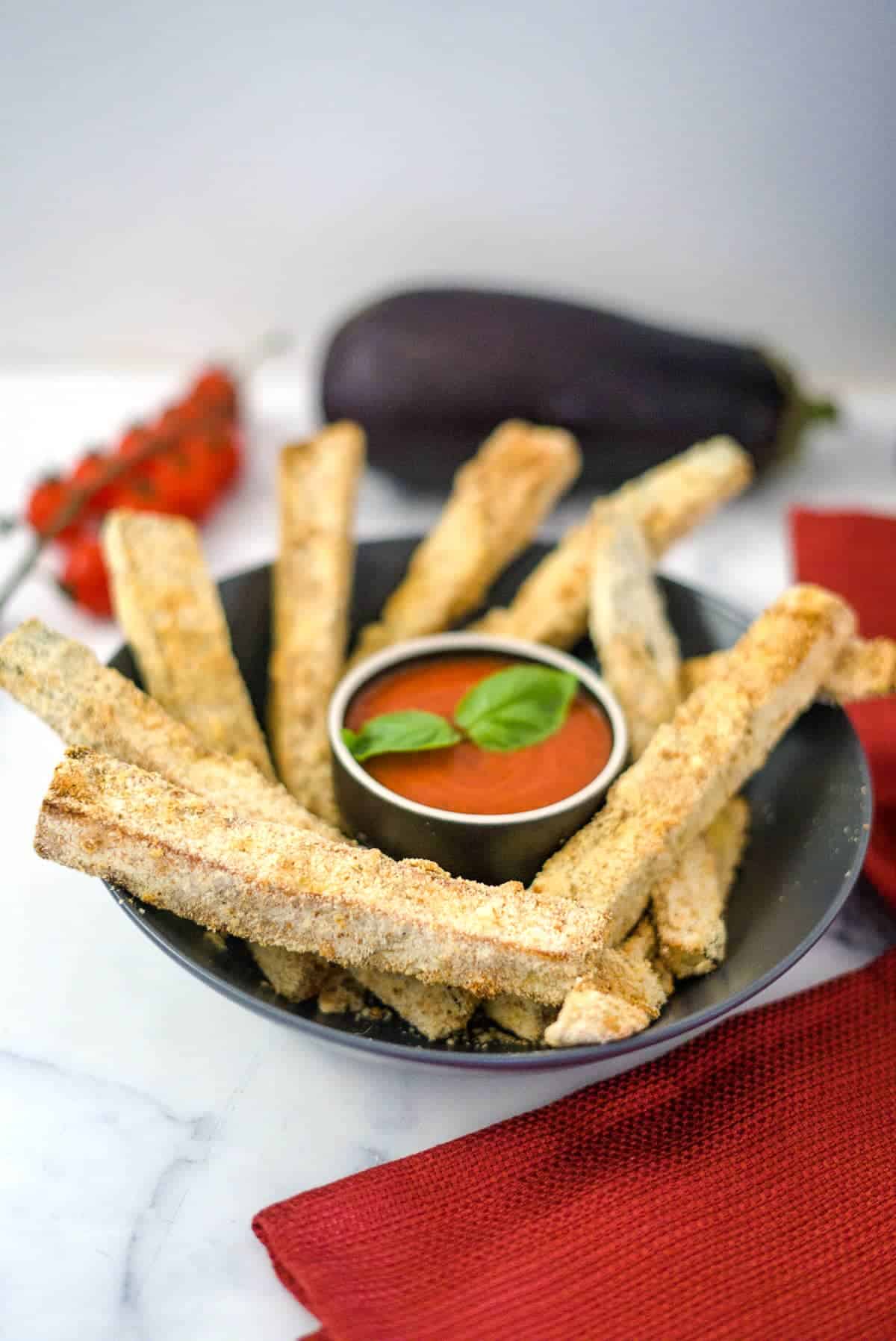 A bowl of crispy, breaded, and baked eggplant fries with a side of marinara sauce, garnished with basil leaves. An eggplant and cherry tomatoes are in the background on a marble surface.