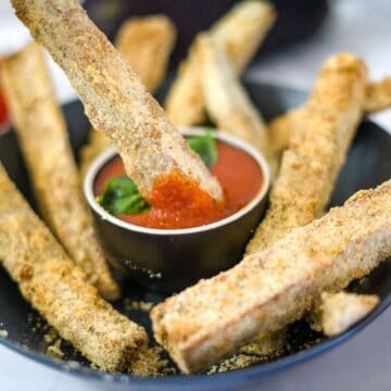 A black bowl contains a mix of breaded zucchini fries and eggplant fries, arranged around a small dish of red dipping sauce, with one zucchini fry partially dipped in the sauce.