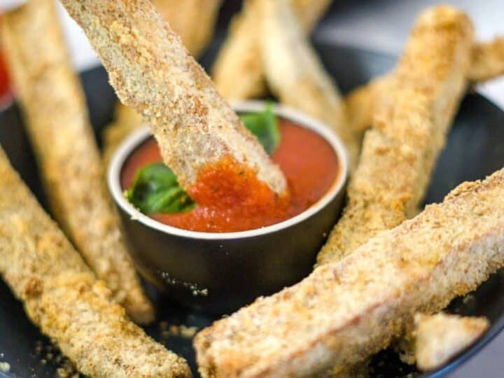 A black bowl contains a mix of breaded zucchini fries and eggplant fries, arranged around a small dish of red dipping sauce, with one zucchini fry partially dipped in the sauce.