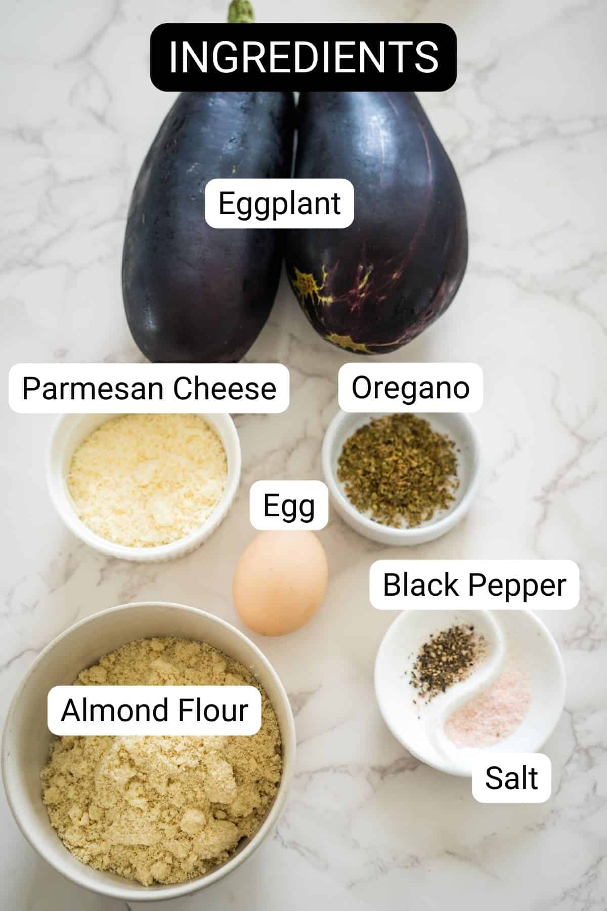 Ingredients displayed on a marble surface: two eggplants ready to be transformed into crispy eggplant fries, a bowl of Parmesan cheese, a bowl of oregano, an egg, a bowl of black pepper, a bowl of salt, and a bowl of almond flour.