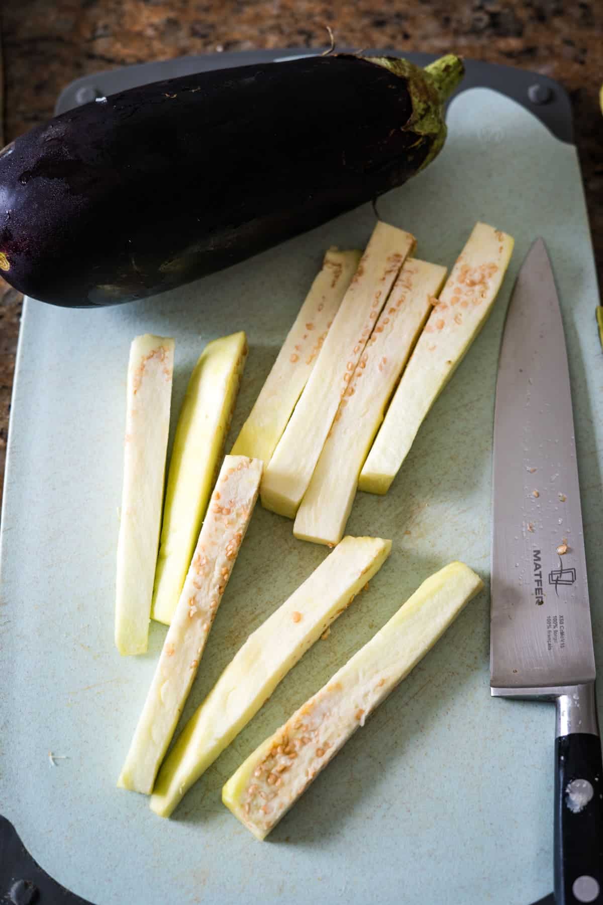 A whole eggplant and several peeled, sliced eggplant pieces are on a cutting board next to a chef's knife, ready to be transformed into delicious eggplant fries.