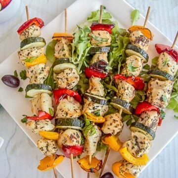 Four grilled chicken and vegetable skewers arranged on a bed of greens on a white plate, featuring succulent chicken, zucchini, bell peppers, and herbs. This healthy chicken recipe is completed with a few olives on the side.