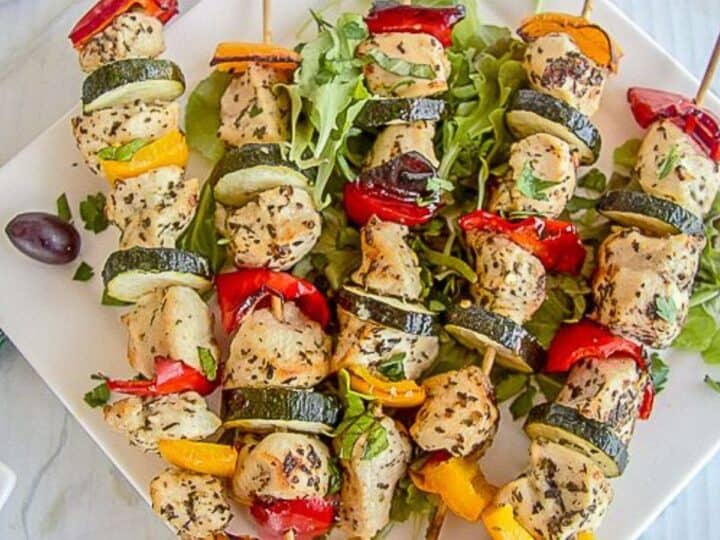 Four grilled chicken and vegetable skewers arranged on a bed of greens on a white plate, featuring succulent chicken, zucchini, bell peppers, and herbs. This healthy chicken recipe is completed with a few olives on the side.