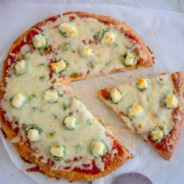 A jalapeno popper pizza with one slice removed, topped with melted cheese, jalapeño slices, and dollops of cream cheese on a parchment-lined surface.