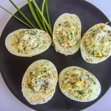 Five keto deviled eggs rest on a black plate, garnished with spices and green herbs, next to fresh chives.