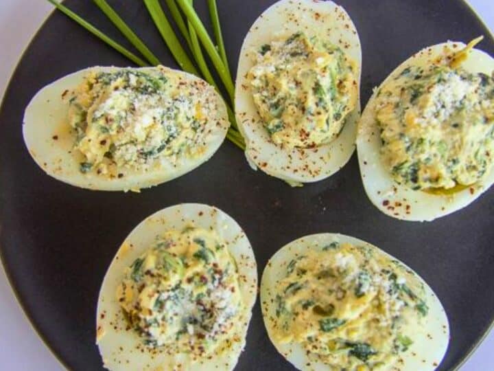 Five keto deviled eggs rest on a black plate, garnished with spices and green herbs, next to fresh chives.