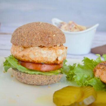 A salmon burger with lettuce and tomato on a whole grain bun, served on a plate with pickles and a side of bbq chicken burgers in a white bowl in the background.