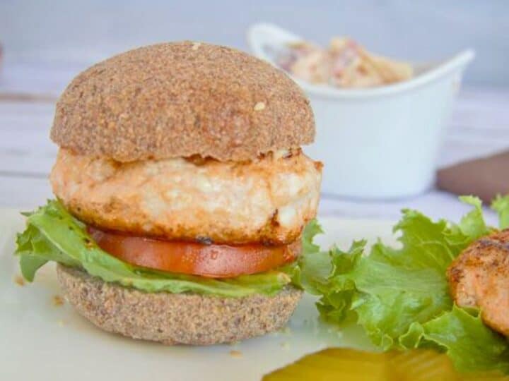 A salmon burger with lettuce and tomato on a whole grain bun, served on a plate with pickles and a side of bbq chicken burgers in a white bowl in the background.