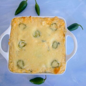 A cauliflower jalapeño popper casserole bakes to perfection in a square dish, with visible jalapeños peeking through. It sits invitingly on a light blue surface, surrounded by whole jalapeños that promise an extra kick.