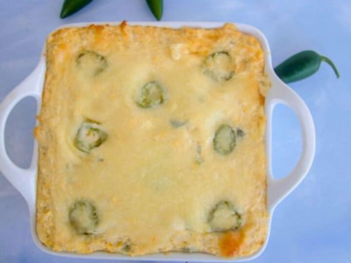 A cauliflower jalapeño popper casserole bakes to perfection in a square dish, with visible jalapeños peeking through. It sits invitingly on a light blue surface, surrounded by whole jalapeños that promise an extra kick.